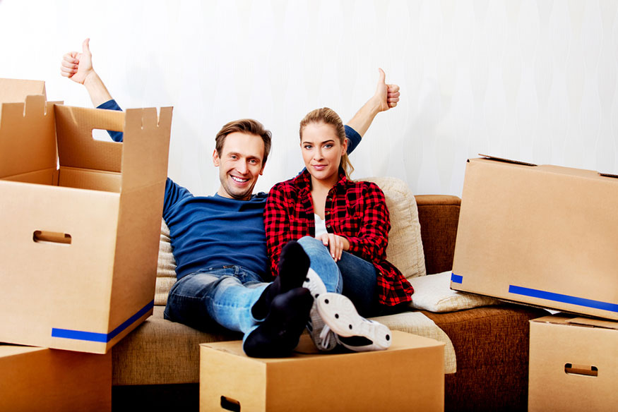 a couple sits on a couch with empty moving boxes, the man has two big thumbs up in triumph